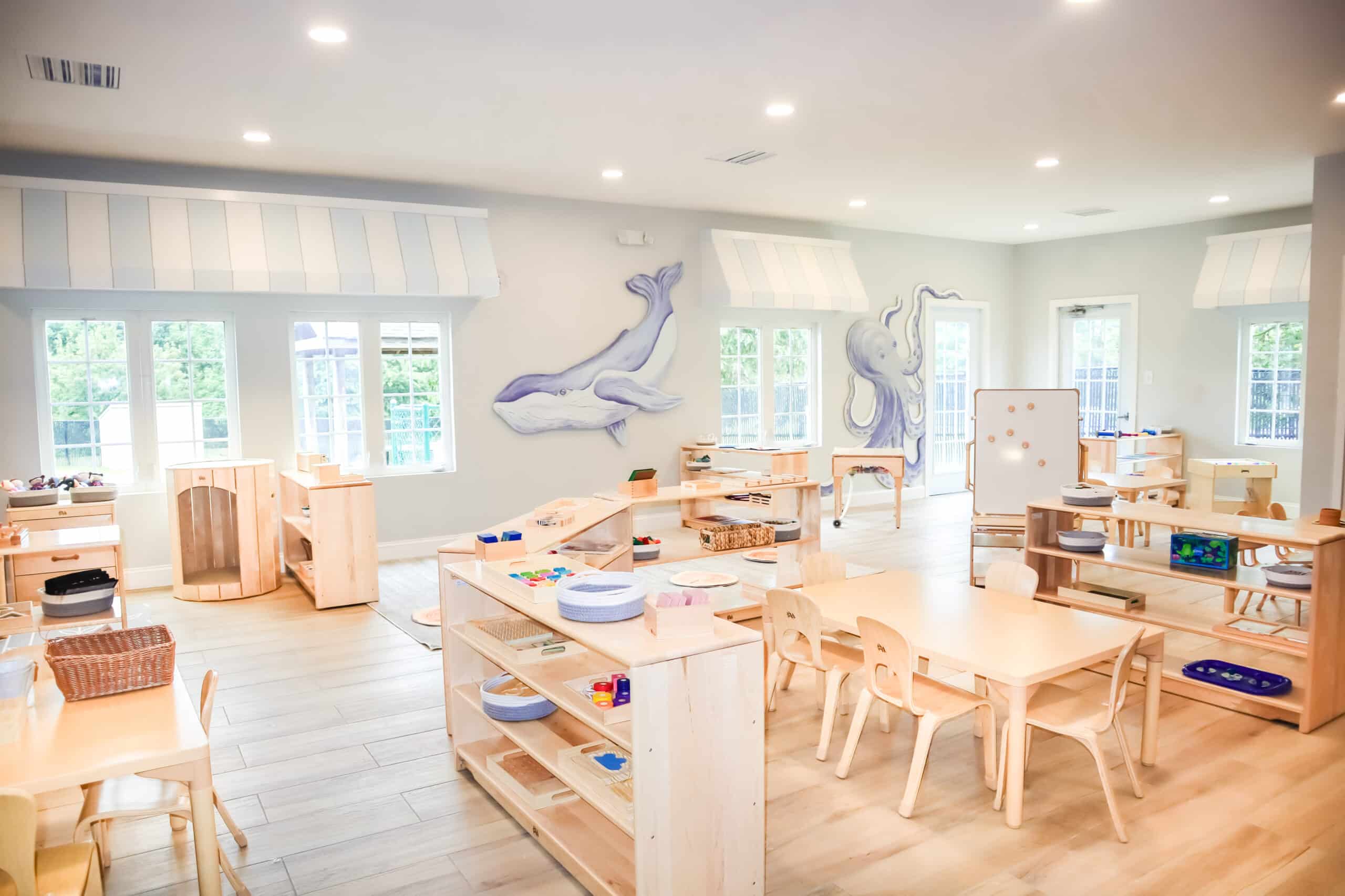 Bright and spacious Montessori-inspired classroom with natural wooden furniture, child-sized tables and chairs, open shelves with learning materials, and ocean-themed wall art featuring a whale and an octopus. Large windows bring in natural light, creating a warm and inviting environment for young children.
